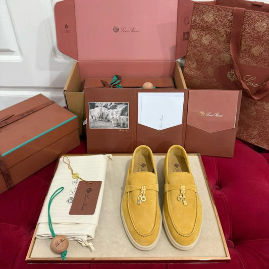 A pair of yellow suede Summer Charms Walk Loafer shoes displayed in front of an open box with various accessories, placed on a red textured surface.
