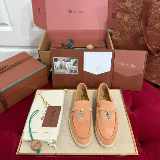A pair of brown suede loafers with decorative charms, displayed in front of a brown box with the brand's logo. A name tag and a key chain are also present, suggesting a package gift or presentation setting.
